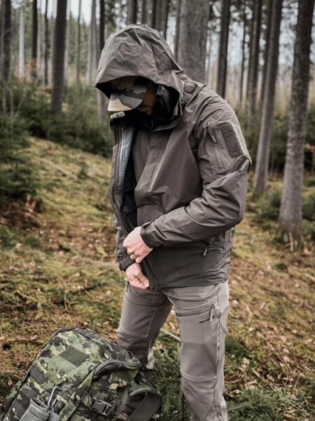 Muž v nepremokavej bunde Helikon-Tex Chinook s kapucňou vedľa maskovacieho batohu v lese.