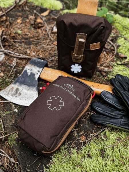 Lekárnička Helikon-Tex Bushcraft Medical Kit v hnedom puzdre, položená v lese pri sekierke a rukaviciach – lekárnička pre bushcraft a outdoor. Foto: Rigad.