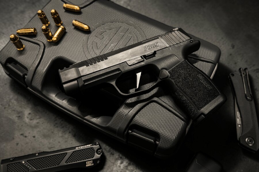 Čierna pištoľ SIG Sauer P365 na prepravnom kufri s logom výrobcu, obklopená nábojmi, zatváracím nožom a svietidlom.